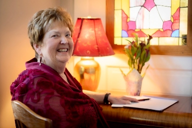 Donna Brown Benton smiling at her writing desk beside a stained-glass window